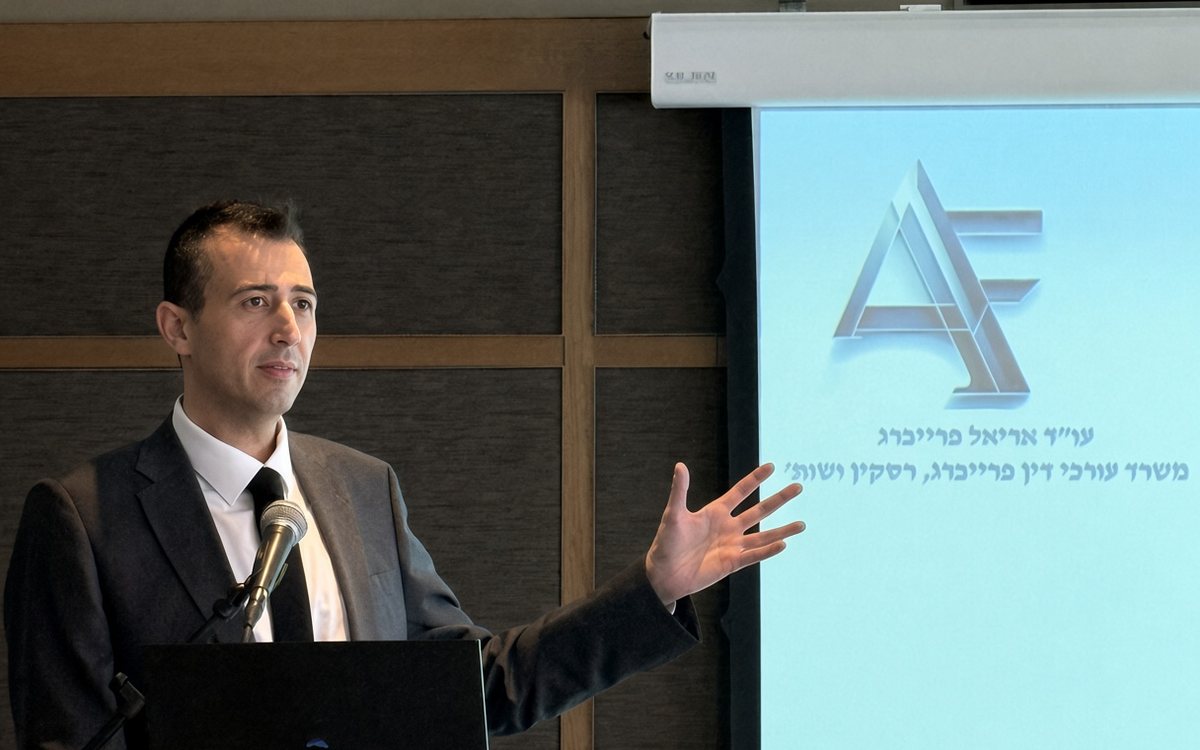 עו״ד אריאל פרייברג מרצה בכנס מקצועי, ולצדו מסך עם לוגו המשרד — משרד עורכי דין אריאל פרייברג ושות׳